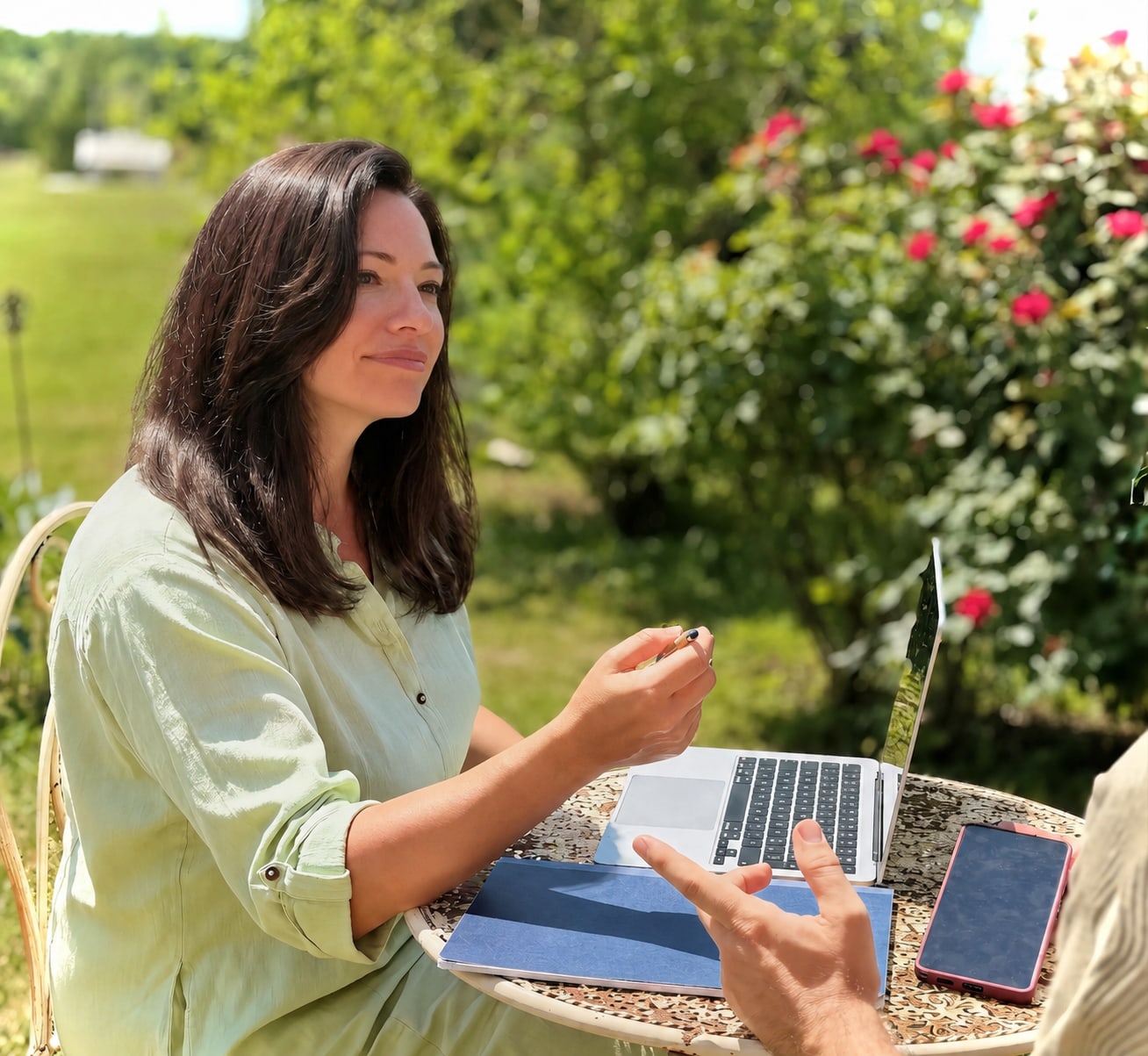 Silvia ascolta un cliente al tavolino in giardino, laptop e quaderno davanti, il roseto rosso alle spalle